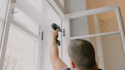 Back view of unrecognizable craftsman using cordless screwdriver and assembling window while screwing metal hinge during renovation works in apartment