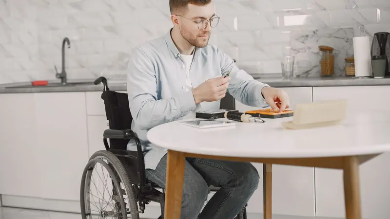 Man in wheelchair fixing electronics on a table in a modern kitchen setting.