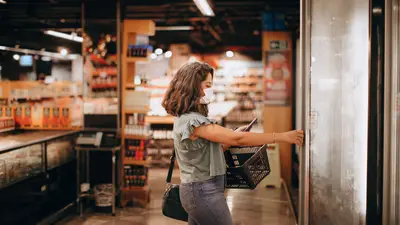 Woman wearing mask shops in grocery store, reaching for freezer door, holding basket.