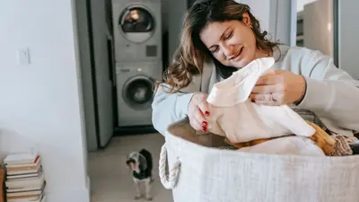Optimistic female with clean clothes in basket having phone conversation while standing in light washroom with washing machines and dog