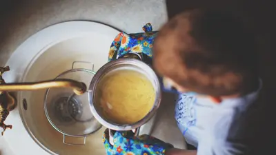 A child strains spaghetti at the sink wearing colorful oven mitts, highlighting a child's culinary exploration.