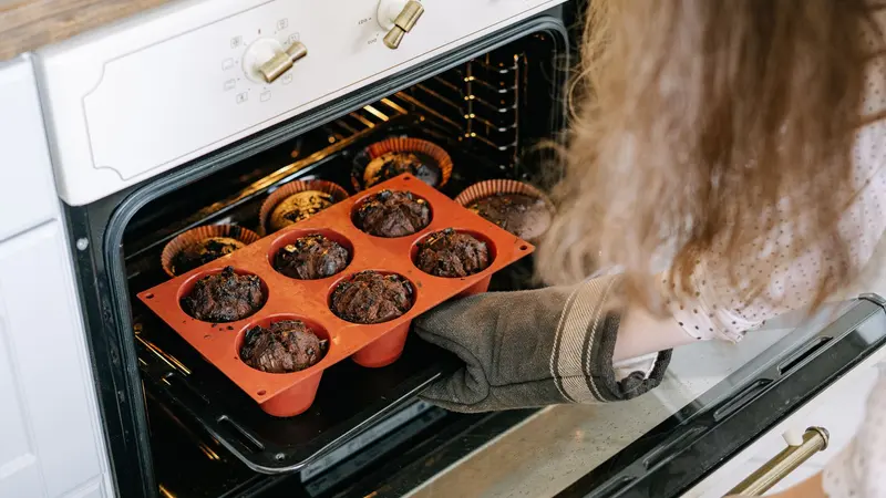 A person taking freshly baked chocolate muffins out of the oven at home.