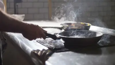 A chef cooks with frying pans on an electric stove in a steamy kitchen.