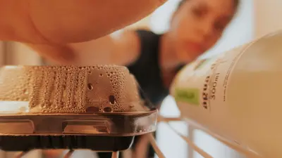 Blurred woman reaching into refrigerator, focusing on food container and drink bottle.